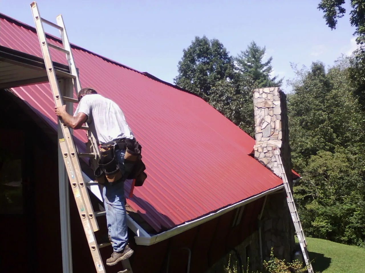 Roofer on a ladder beside a red metal roof and stone chimney — Soffit & Fascia Repair by Roofing Brothers in Santa Paula